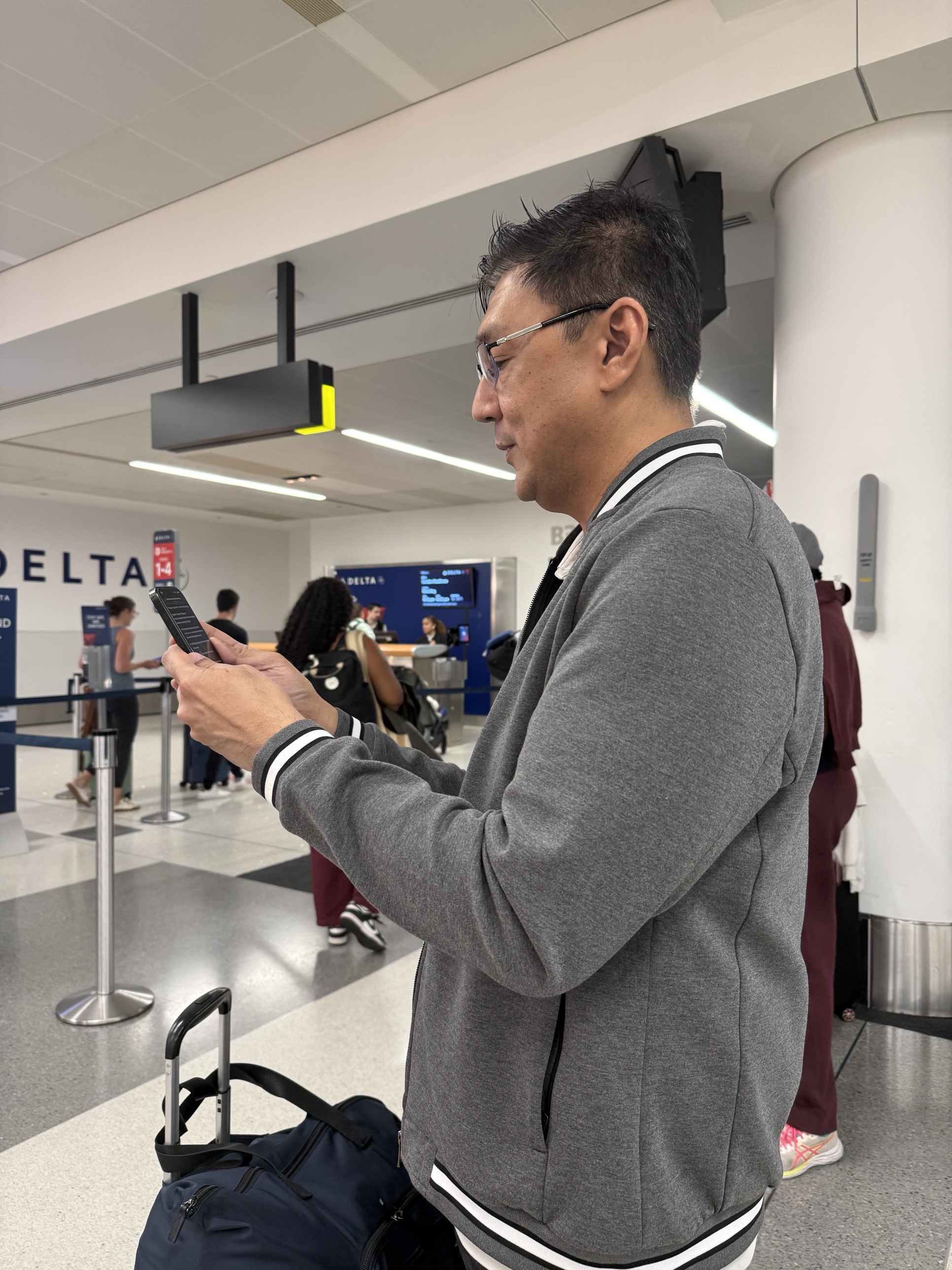 Angelo standing in a Delta boarding area in a grey zip jacket, reading his phone with a carry-on bag at his feet, other passengers and a gate counter in the background.
