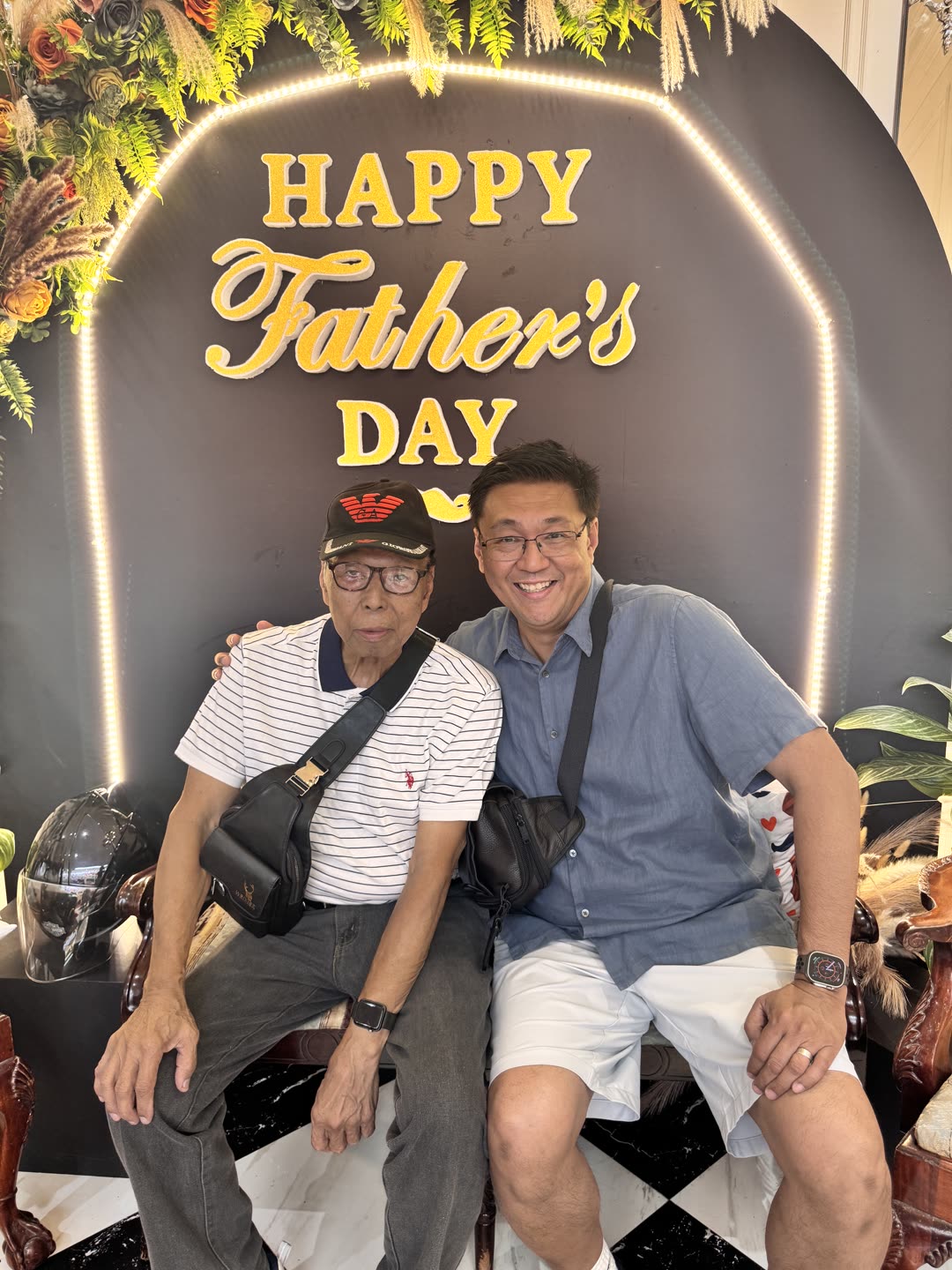 Angelo and his father sitting together in front of a Happy Father's Day backdrop, both smiling, surrounded by warm lights and flowers.